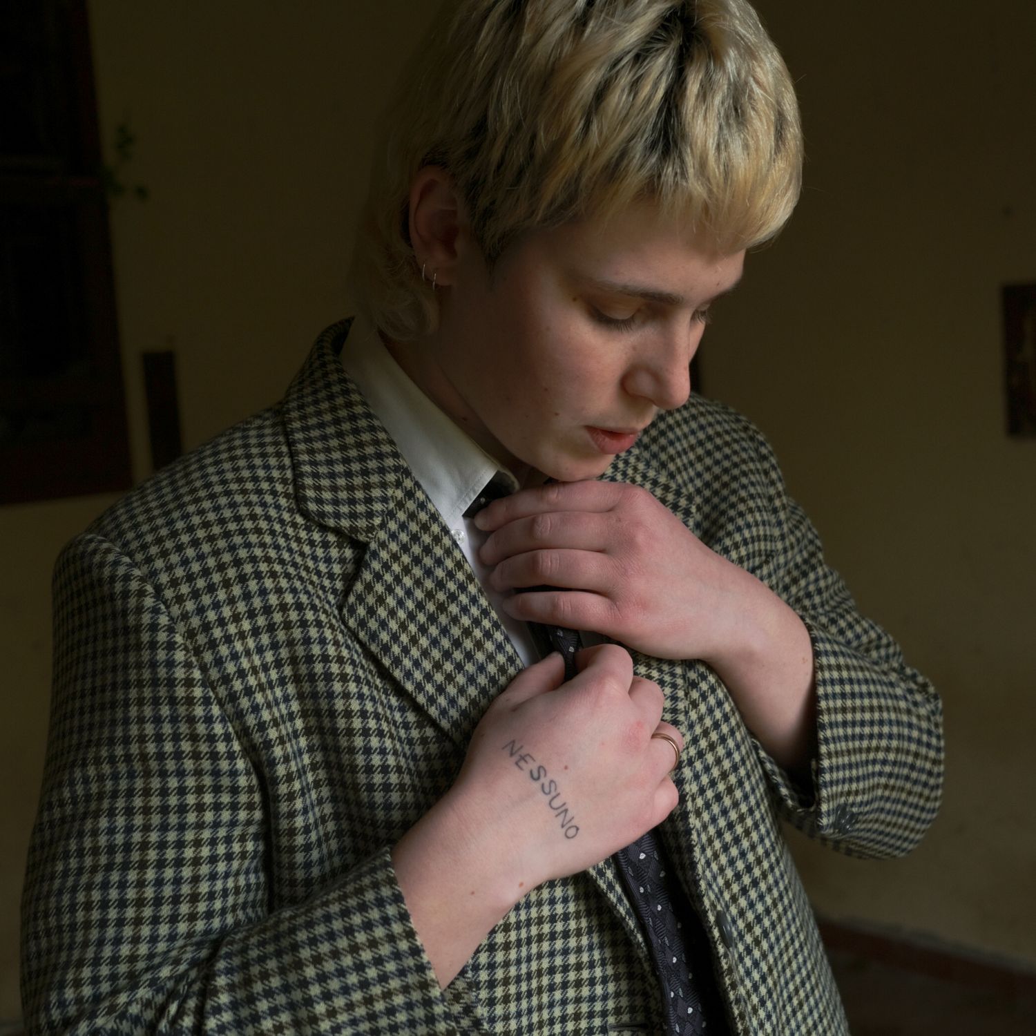 A blond person in a checked blazer looks down, hand on their lapel